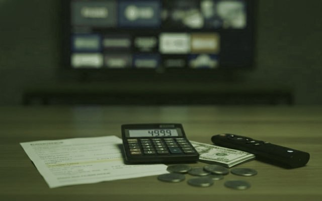 Calculatrice, billets, pièces, clé de streaming et télécommande sur une table devant un téléviseur